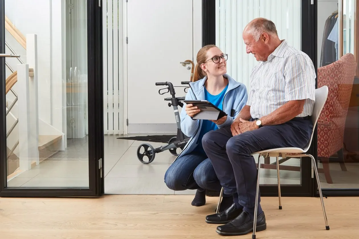 A care worker with a tablet kneels beside an older client at home