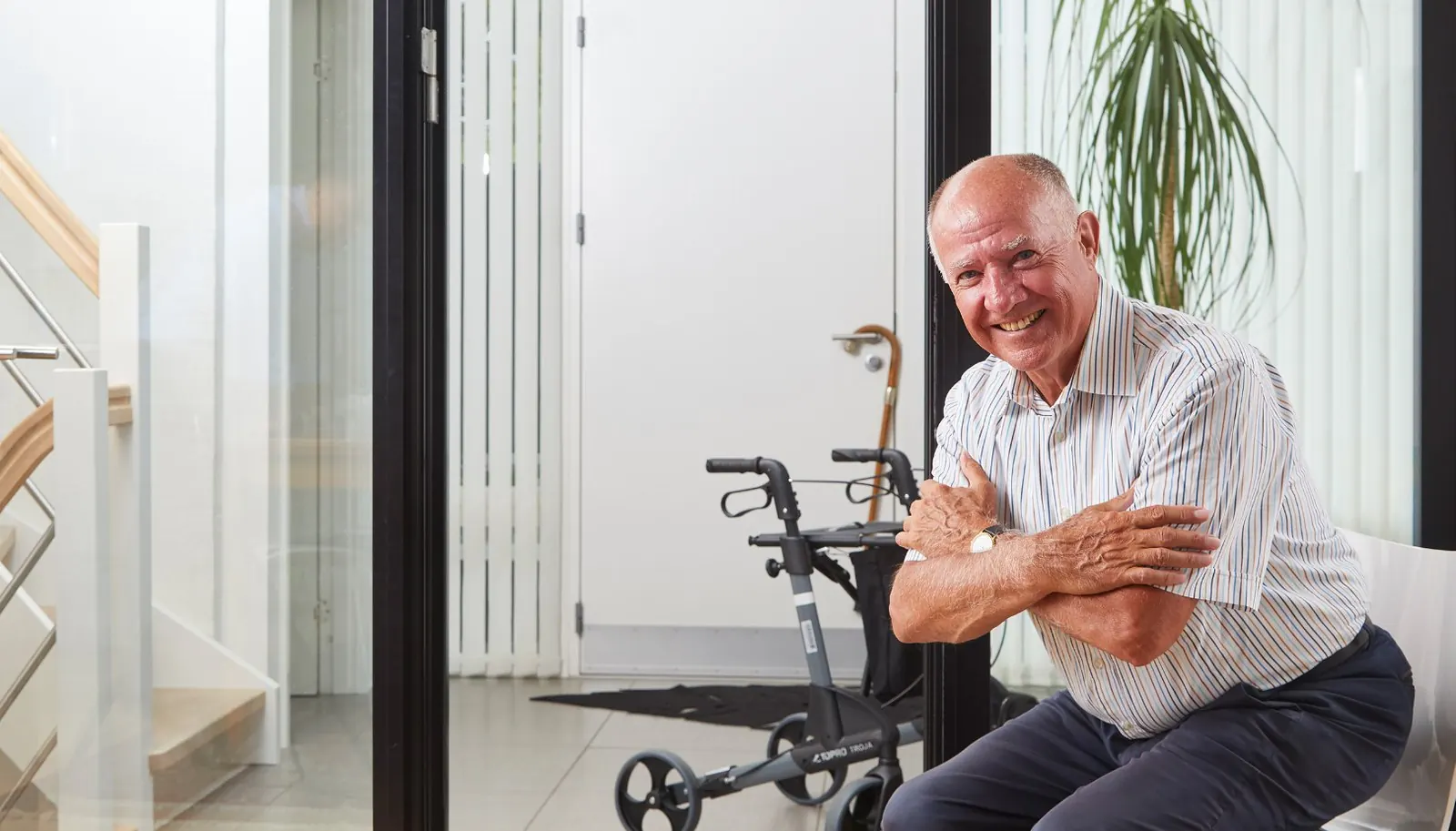 An older man sits smiling at home, his rollator behind him