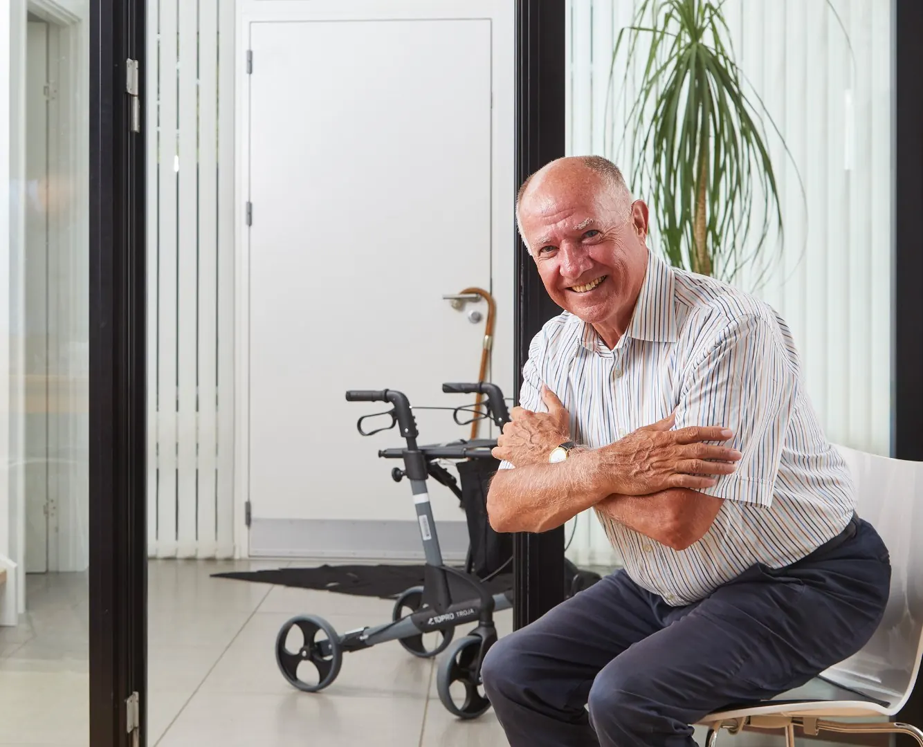 An older man sits smiling at home, his rollator behind him